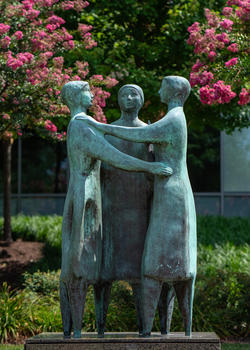 Copper statue titled Communitas, green from oxidation. It shows three feminine figures standing in a circle, arms around each other.